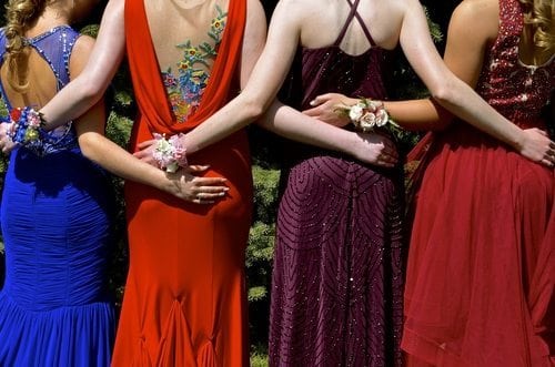 four young women wearing prom dresses with their arms around each other, photo taken from behind