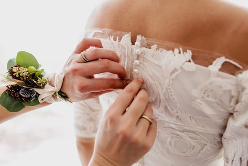 Hands buttoning up a wedding dress with lace details and pearl buttons.