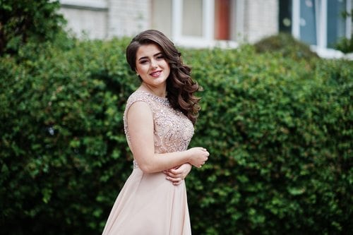 A girl in a pale pink prom dress posing in front of a green bush.